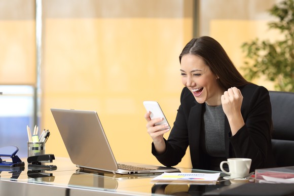 A businesswoman in front of an open laptop looking at phone and cheering