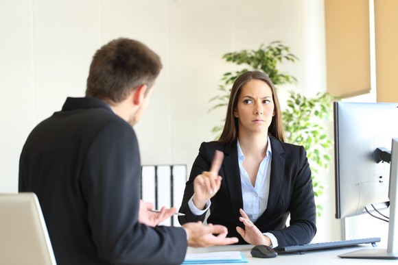 A woman with a stern look on her face holds a finger up to a man sitting across from her