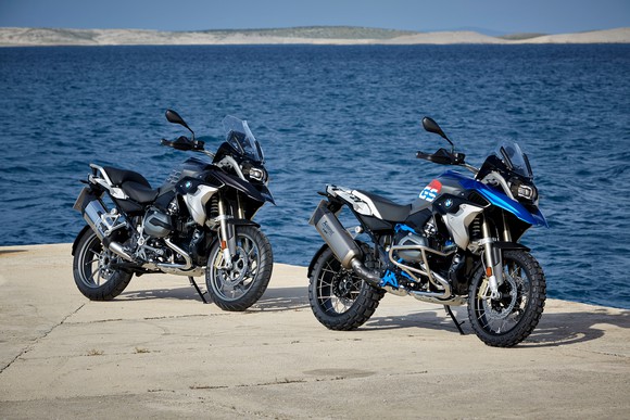Two BMW R 1200 GS motorcycles, in black with blue trim, parked on a sunny waterfront.