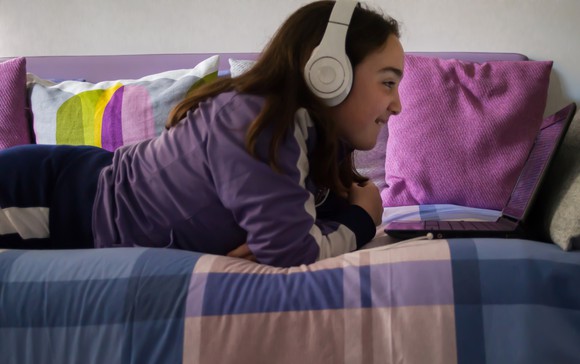 Girl laying on daybed smiling, wearing headphones watching laptop.