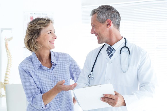 Middle-aged female patient smiling looking at face of smiling male physician who has a stethoscope around his neck and is holding papers.