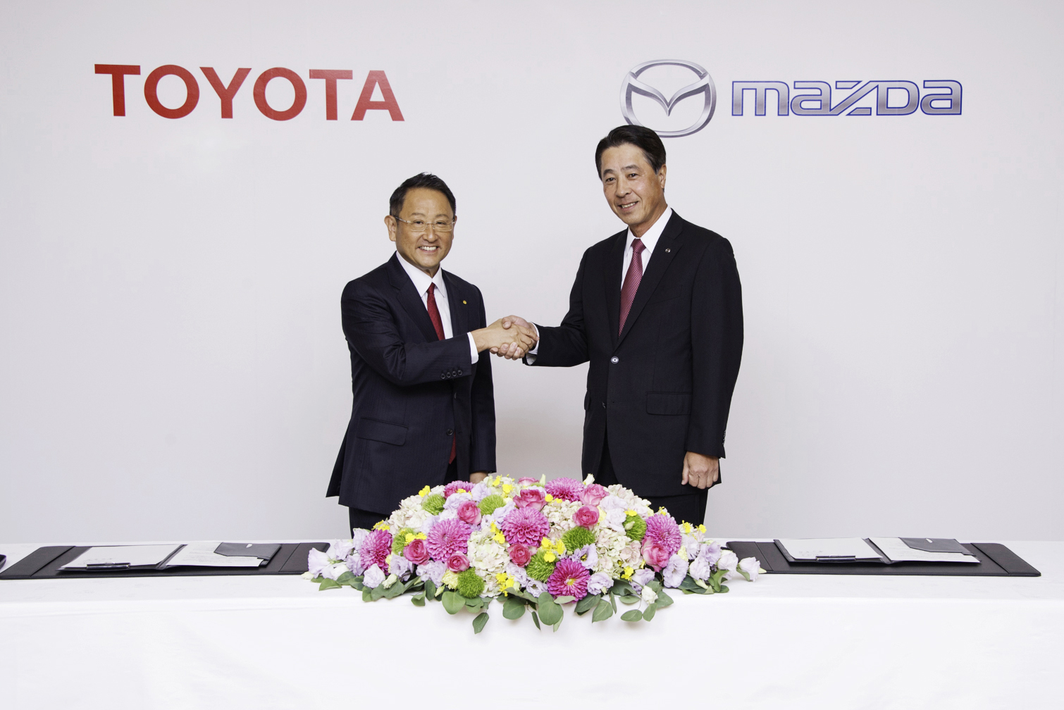 Akio Toyoda and Masamichi Kogai are shown shaking hands before a backdrop with the Toyota and Mazda logos.