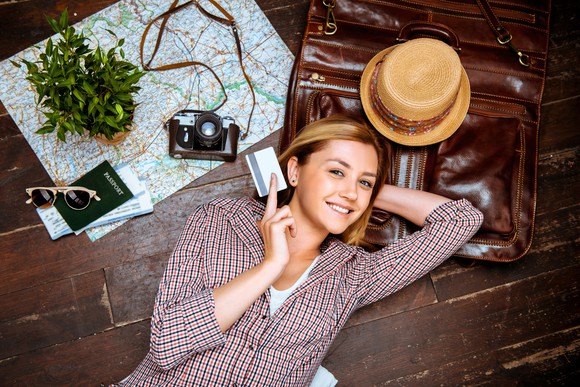 A downward-looking view of a smiling woman holding a credit card, lying on a floor with a suitcase, hat, map, camera, and passport.