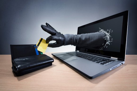 A gloved hand reaching through a computer screen to steal a bank card from a wallet.