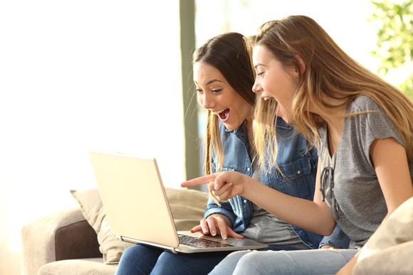 Two young women smiling at something on their laptop screen. One is pointing at the screen.