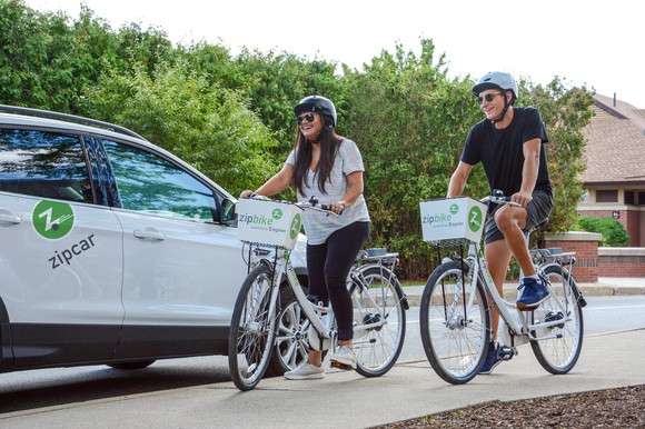People riding bicycles next to a Zipcar.