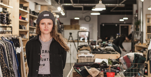 A young woman business owner dressed casually with knit cap in her rustic looking store of clothes that match what she's wearing.