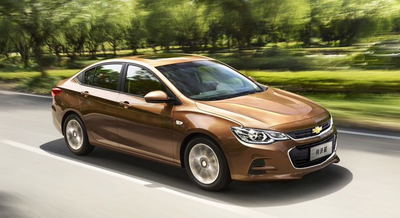 A brown 2017 Chevrolet Cavalier with a Chinese-language license plate, driving on a road near a wooded field.
