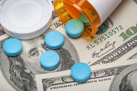 Blue pills spill out of an overturned prescription bottle and onto a pile of hundred-dollar bills.
