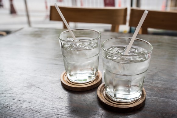 Two glasses of cold water with straws in them sitting on coasters on a wooden table.