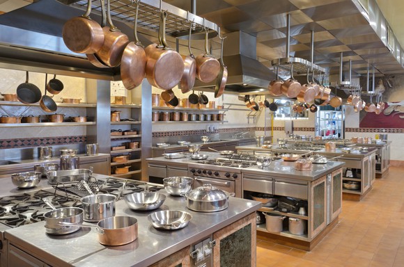 Professional kitchen with stoves, pots, pans, and other kitchen tools on three large tables and several racks.