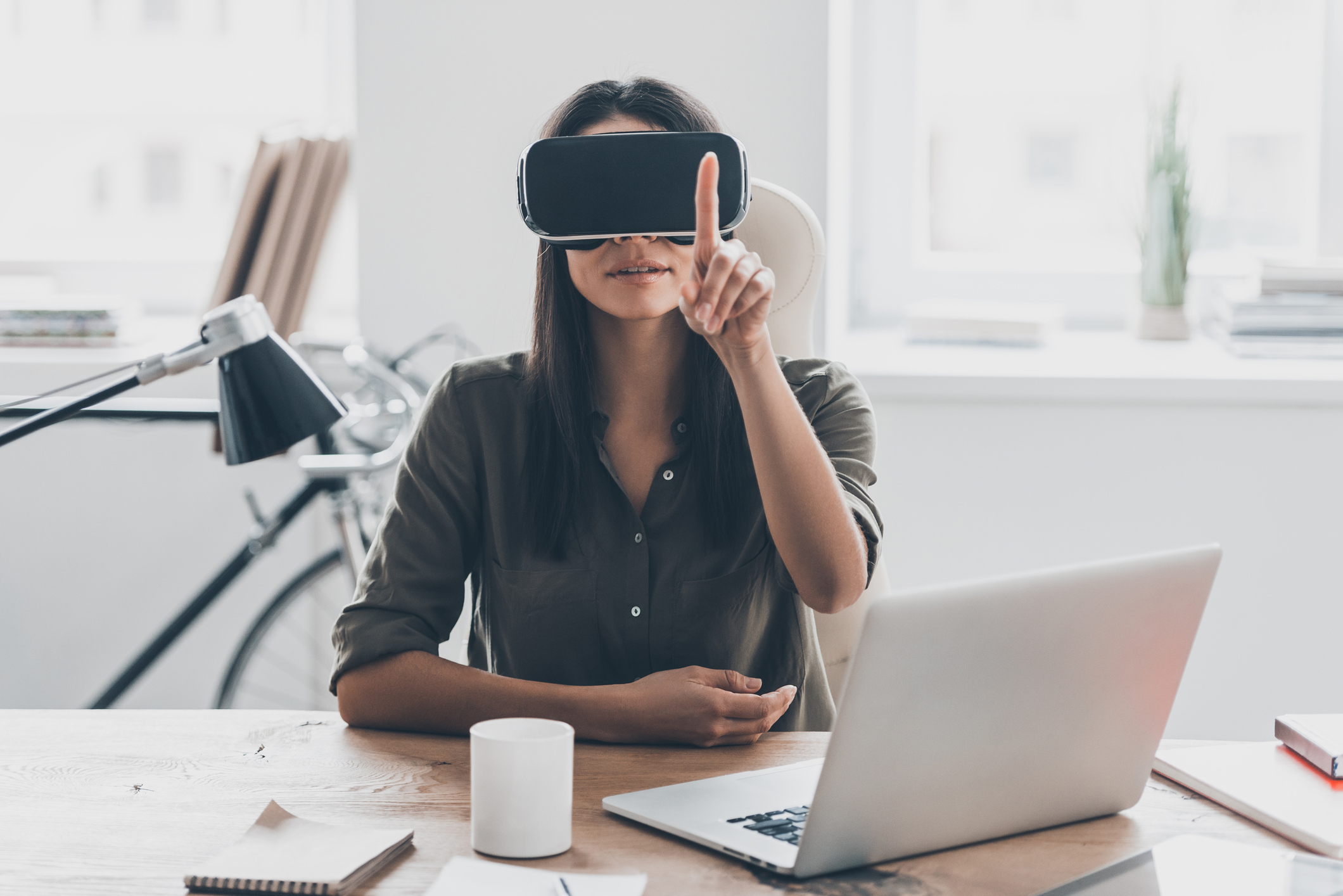 Woman sitting at desk wearing a virtual reality head and pointing her finger in the air.