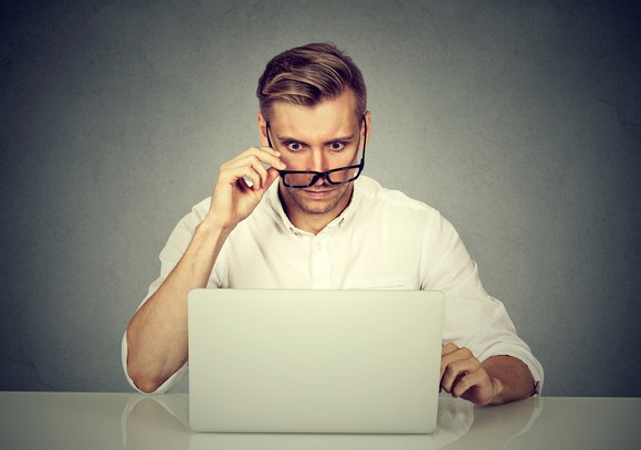 A man at a desk staring at his laptop, pulling his glasses down as he's surprised about what's on the screen.