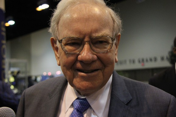 Warren Buffett wearing a gray suit, blue tie, and smiling into the camera