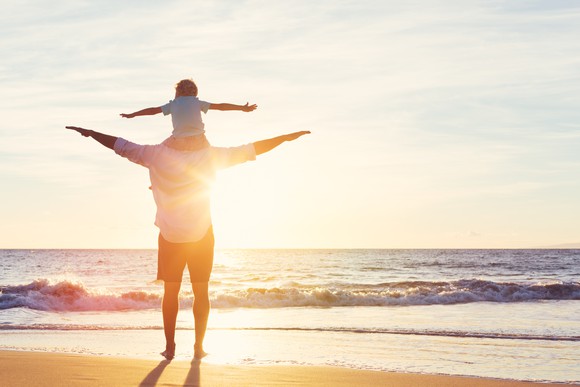 With a child on his shoulders, a man looks at the sun setting over the ocean from the shoreline.