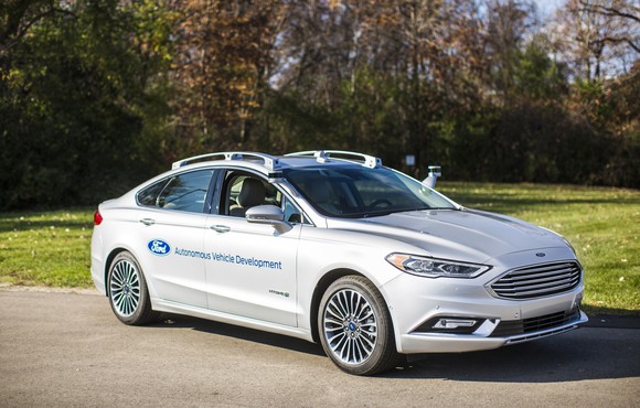 A white 2017 Ford Fusion with self-driving sensors visible, parked near a wooded field.