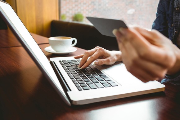 A woman holding a credit card in her left hand while her right hand is on the keyboard of her laptop. There is a cup of coffee on the table.