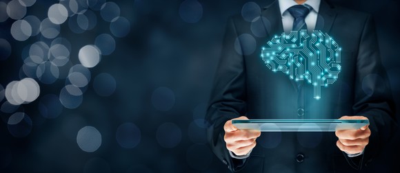 Man in a suit holding a flat, glass tablet above which is the illustration of a lit-up brain.