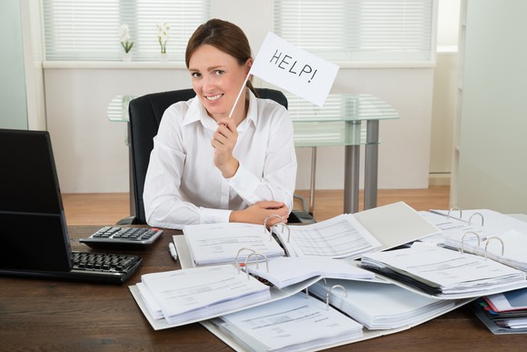 Smiling woman at desk covered with paper, holding up small "help" sign.