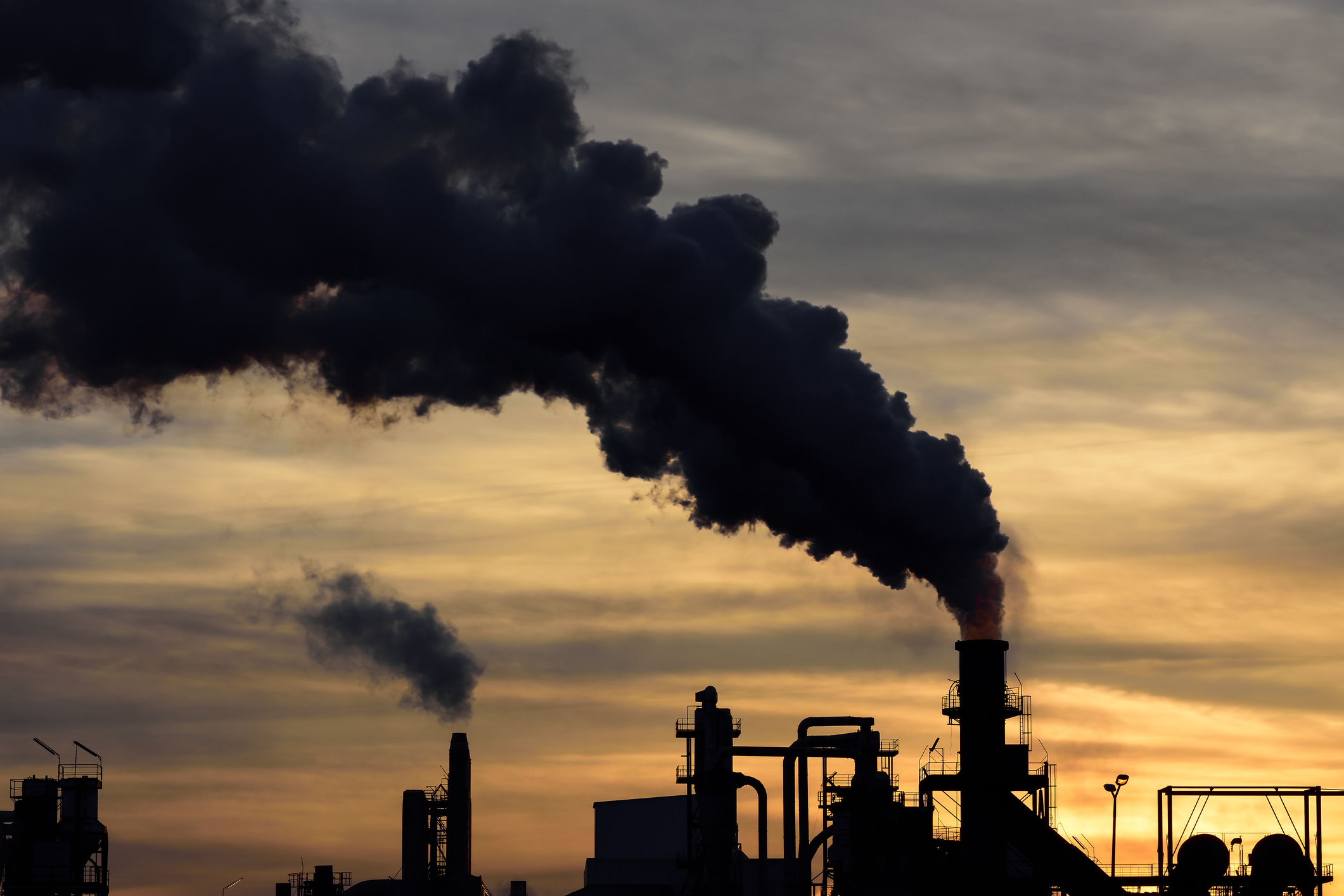 Black smoke billowing out of a factory smokestack silhouetted against a sky at sunset