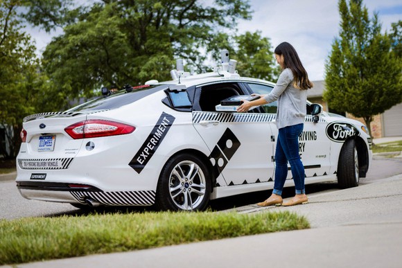 A woman takes pizzas from a self-driving Ford Fusion Hybrid pizza-delivery test vehicle at a curb in front of a house.