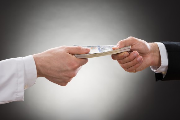 Hand in white sleeve holding money on one end with hand in dress shirt and coat holding other end