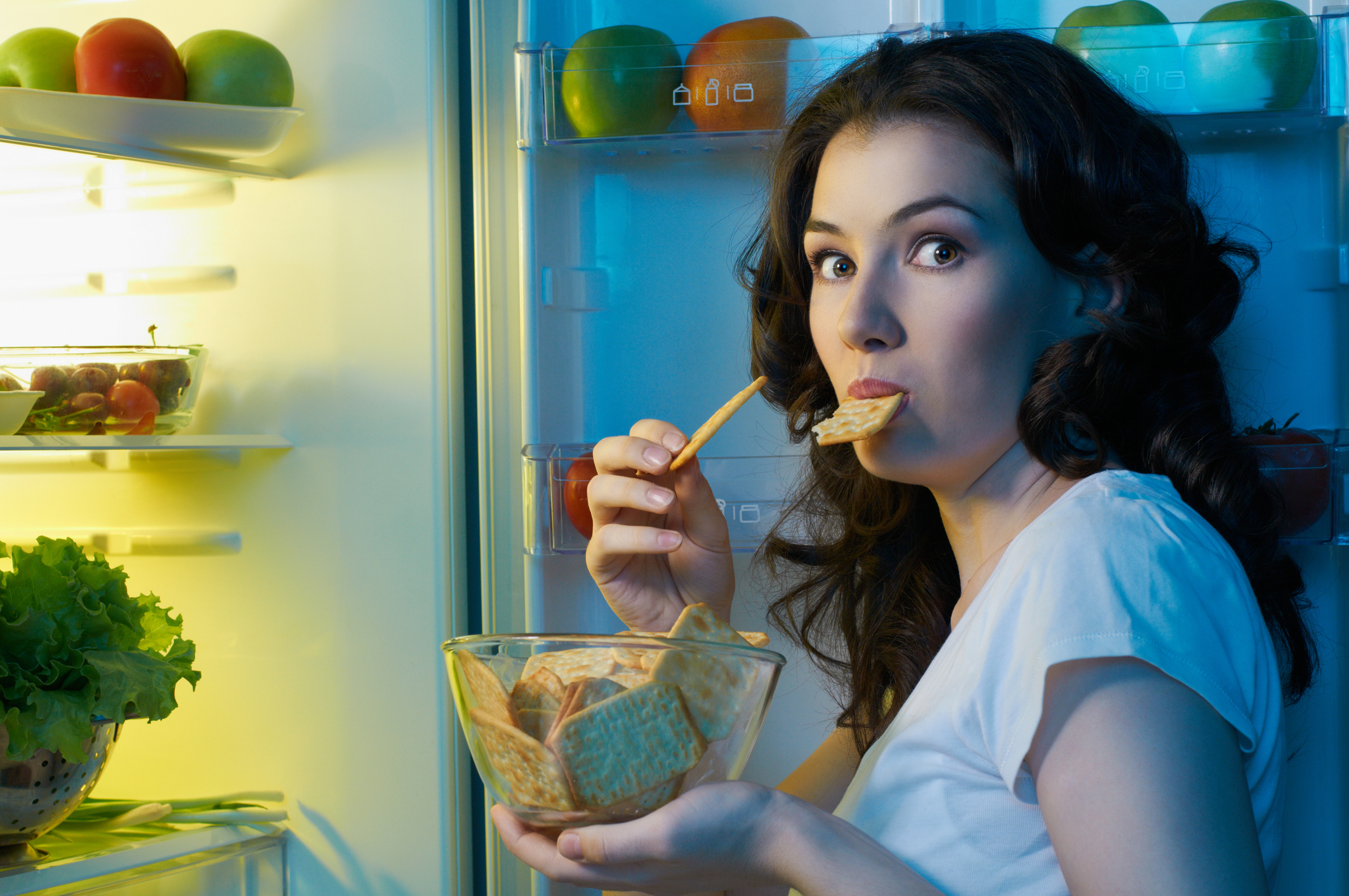 Woman holding a bowl of crackers, one in her mouth, standing at a refrigerator looking surprised like someone who has been caught red-handed.