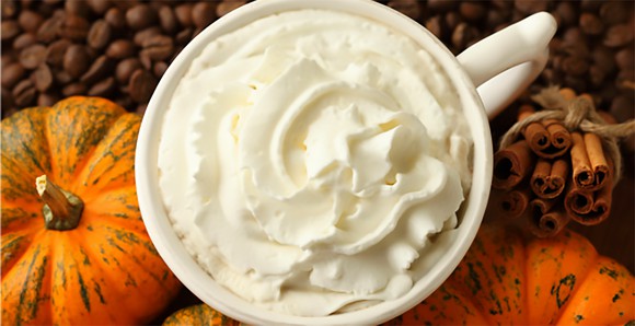 An overhead shot of a Starbucks Pumpkin Spice Latte, showing the whipped cream topping. Gourds, coffee beans, and cinnamon sticks are also on the table.
