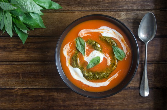 Bowl of tomato soup on wooden table, with swirls of pesto and sour cream on surface, sprinkled with fresh basil leaves