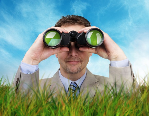 Man dressed in suit standing above tall grass looking through binoculars