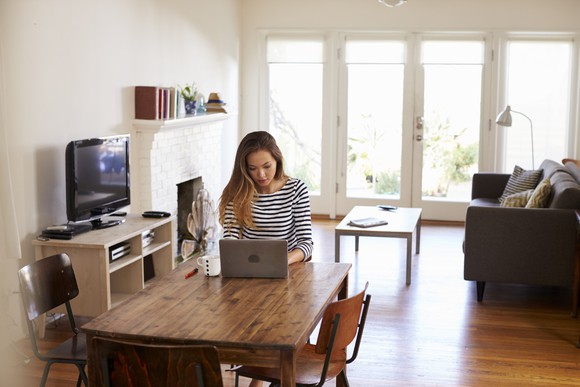 A woman sits at a table in her home, working on a laptop.