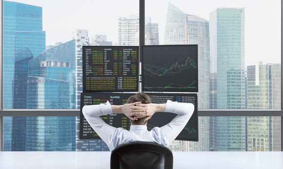 A man sitting in a chair folds his hands behind his head, as he looks at stock-related information on a cluster of monitors in front of a window that looks out on a city skyline.