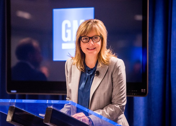 Mary Barra is shown standing at a podium in front of a lighted GM logo. 