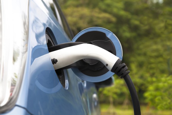 Blue electric car with charging cord in it. Greenery background.