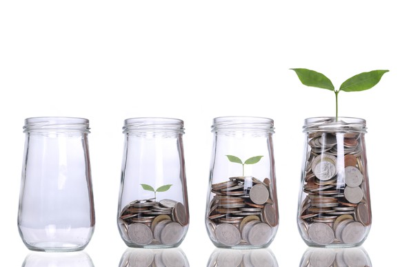 A row of jars that are increasingly full of coins, with a plant growing out of the fullest jar.