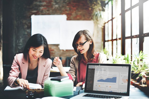 Two businesswomen seated at a table with a laptop facing outward, showing data visualizations