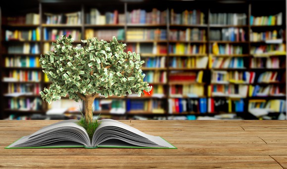 Tree with stylized dollar-bill leaves growing out of an open book, on a table in a library