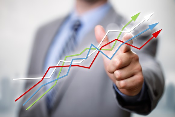 A man in a suit points to a grouping of lines -- green, white, blue, and red -- going upward, as if following a stock chart.