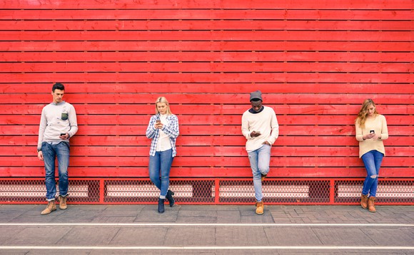 Four young people stand against a red wall, using cellphones.