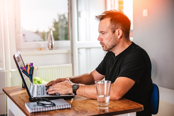 A man types on a laptop as he sits by a sunny window. Near him are a pad of paper, a drinking glass, reading glasses, and a container of colorful pens.