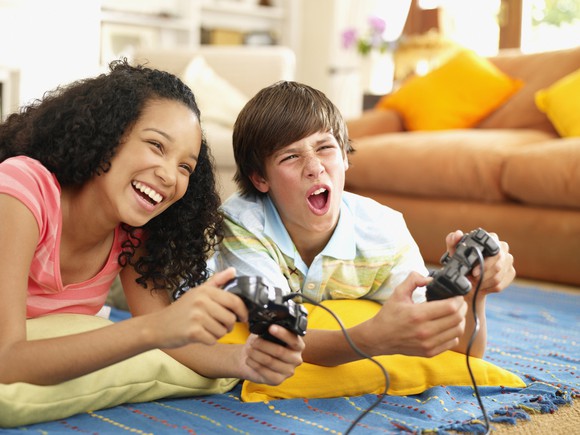 Two kids play a console game while lying on the floor.