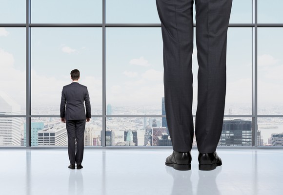 Small man next to giant man looking through window at city