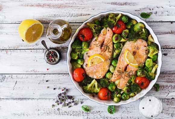 Bowl of colorful salad with salmon steaks on top, next to half a lemon, a bottle of oil, a small bowl of salt, and a container of peppercorns on a whitewashed table