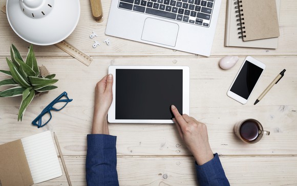 Hands use a tablet while a laptop and smartphone are nearby.