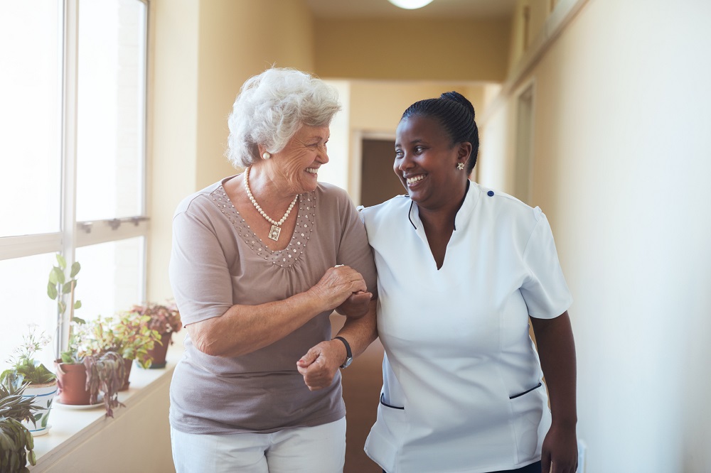 Senior woman and care attendant walk hand in hand and smile at one another.