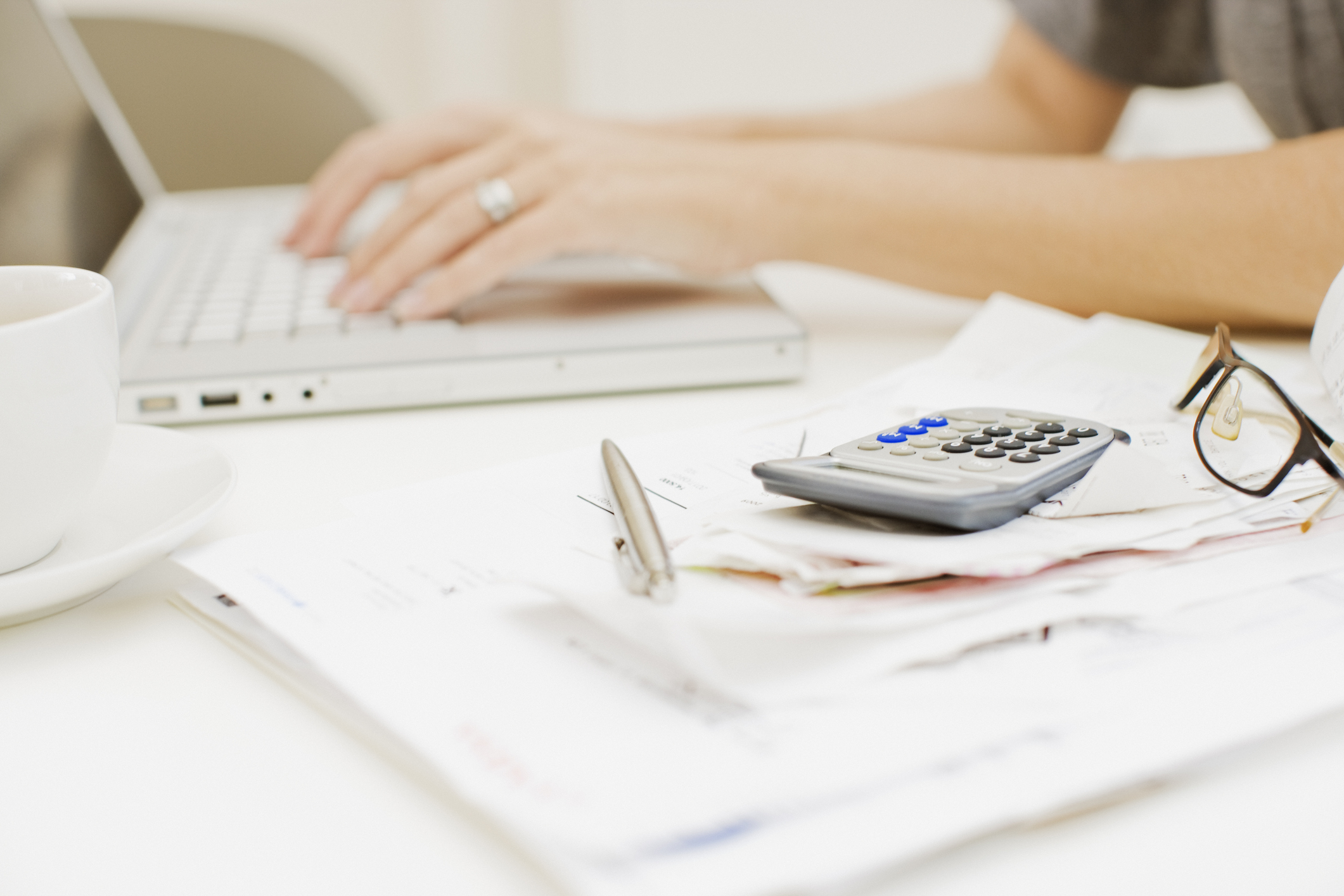 Person typing on a laptop with papers, a pen, glasses, and a calculator on hand