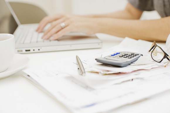 Person typing on a laptop with papers, a pen, glasses, and a calculator on hand
