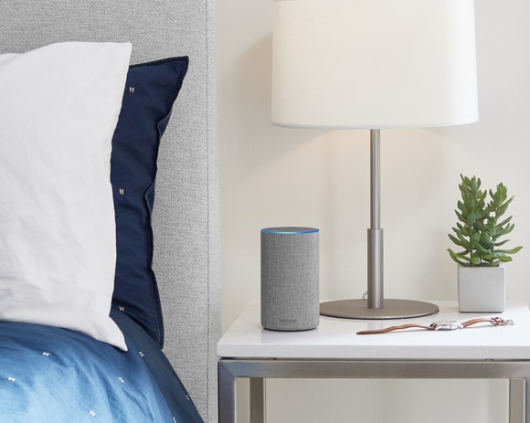 The new Amazon Echo speaker on a table next to a bed.