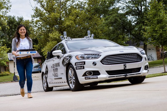 A Ford Fusion with visible self-driving sensors is in a suburban driveway. A woman holding a boxed pizza is walking away from the car.