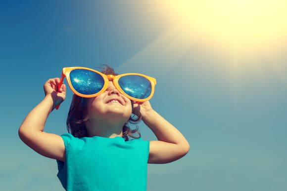 Smiling little girl wearing big sunglasses looks upward with sun in background